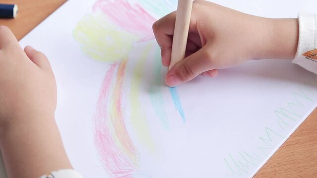 A preschooler draws with colored pencils in an album. Child's hand with a pencil close-up, selective focus. The girl draws a rainbow