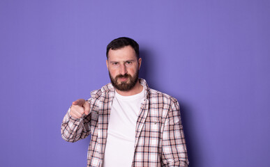 Obraz premium Portrait of angry bearded handsome young man pointing index finger at you on purple isolated background, looking at camera.
