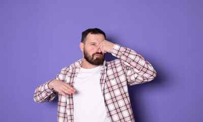 Bad smell. Young bearded man on purple background wearing casual smelling something stinky and disgusting, intolerable smell, holding breath with fingers on nose.