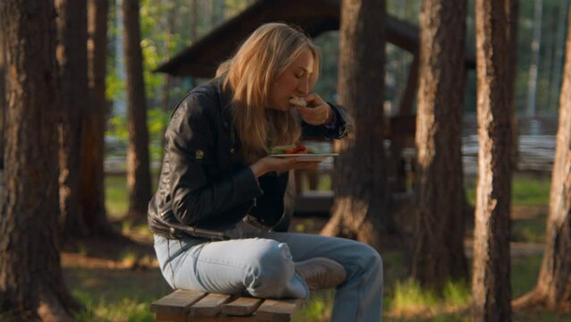 Beautiful young woman eating alone in park. Stock footage. Attractive young woman eats on bench in forest recreation center. Woman eats alone in park on sunny summer day