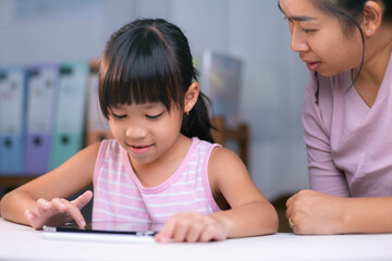 Happy little Asian daughter talking with mother using digital tablet to do homework or study online. Happy young girl and adult sitting at table learning on iPad together. Back to school.
