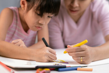 Mother and daughter drawing together with crayons. Adult woman helps girl study or draw together at home in living room. Happy family.