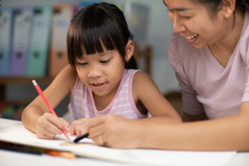 Mother and daughter drawing together with crayons. Adult woman helps girl study or draw together at home in living room. Happy family.