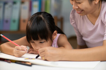 Mother and daughter drawing together with crayons. Adult woman helps girl study or draw together at home in living room. Happy family.