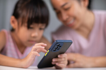 Happy Asian family mother and cute daughter using phone together at home. Mother and little girl laughing happily using smartphone watching funny social media video app sitting at table together.