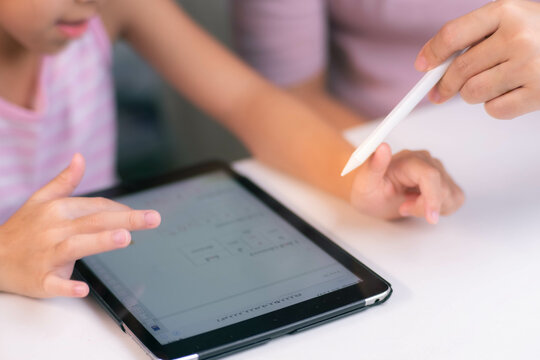 Happy little Asian daughter talking with mother using digital tablet to do homework or study online. Happy young girl and adult sitting at table learning on iPad together. Back to school.