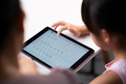 Happy little Asian daughter talking with mother using digital tablet to do homework or study online. Happy young girl and adult sitting at table learning on iPad together. Back to school.