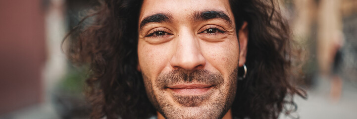 Fototapeta premium Young attractive italian guy with long curly hair and stubble looks into the camera and smiles at old buildings background. Stylish man with an earring in his ear and lot of chains