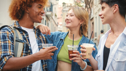 Company of happy friends, two guys and girl, walking with takeout drinks along city street, chatting and laughing