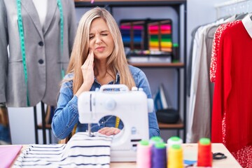 Blonde woman dressmaker designer using sew machine touching mouth with hand with painful expression because of toothache or dental illness on teeth. dentist concept.
