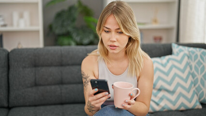 Young blonde woman using smartphone drinking coffee at home