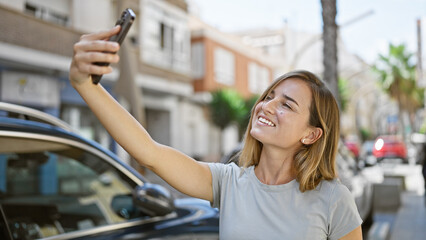 Joyful young blonde woman confidently making a fun selfie with her smartphone on the city street, basking in sunlight while enjoying her urban lifestyle.