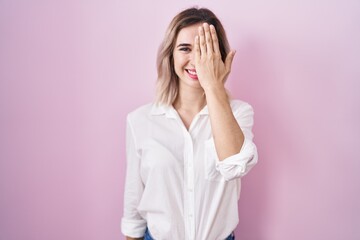 Young beautiful woman standing over pink background covering one eye with hand, confident smile on face and surprise emotion.