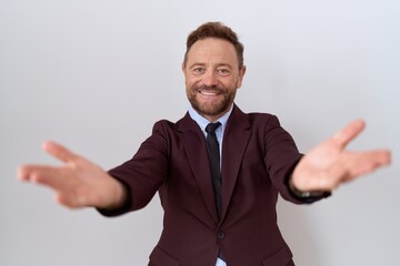 Middle age business man with beard wearing suit and tie looking at the camera smiling with open arms for hug. cheerful expression embracing happiness.