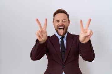 Middle age business man with beard wearing suit and tie smiling with tongue out showing fingers of both hands doing victory sign. number two.