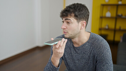 Young hispanic man sending voice message with smartphone sitting on sofa at home