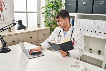Young hispanic man doctor using laptop reading book at clinic
