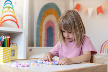 A cute girl is sitting at a table in the children's room and making crafts out of pompoms. Children's crafts.