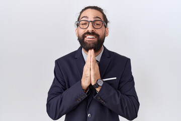 Hispanic man with beard wearing suit and tie praying with hands together asking for forgiveness smiling confident.