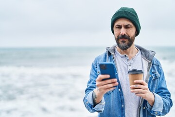 Young bald man using smartphone drinking coffee at seaside