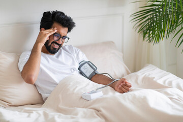 Hypertension. Indian Man Checking Blood Pressure With Upper Arm Monitor In Bed