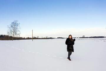 a cheerful girl in a warm fur coat walks across the open field leaving footpath in the snow