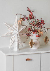 Christmas composition on a white chest of drawers - a Christmas bouquet with cranberry branches and paper decorations, a Christmas star and a ceramic Christmas tree on a white background