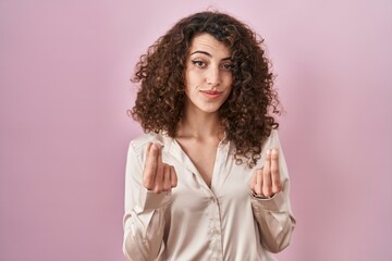 Hispanic woman with curly hair standing over pink background doing money gesture with hands, asking...