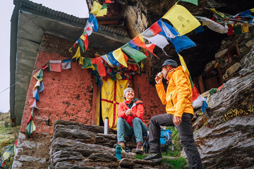 Chatting smiling Backpackers Couple tea break at small sacred Buddhist monastery decorated multicolored Tibetan prayer flags with mantras. Climbing Mera peak route in Makalu Barun National Park, Nepal - obrazy, fototapety, plakaty