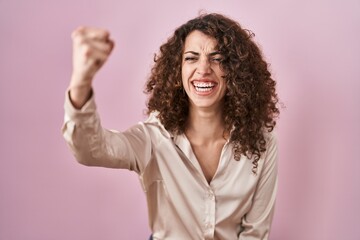 Hispanic woman with curly hair standing over pink background angry and mad raising fist frustrated and furious while shouting with anger. rage and aggressive concept.