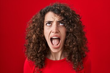 Hispanic woman with curly hair standing over red background angry and mad screaming frustrated and furious, shouting with anger looking up.