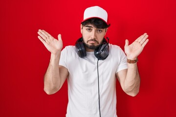 Hispanic man with beard wearing gamer hat and headphones clueless and confused expression with arms and hands raised. doubt concept. © Krakenimages.com