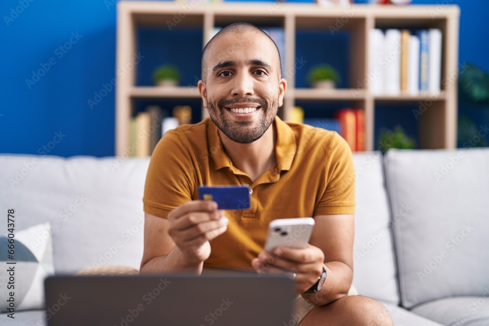 Sticker Young latin man using smartphone and credit card sitting on sofa at home