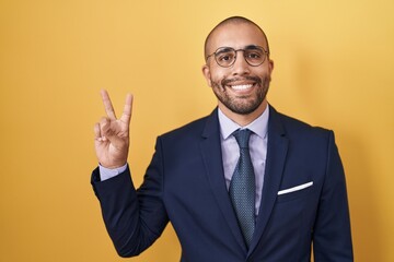 Hispanic man with beard wearing suit and tie showing and pointing up with fingers number two while smiling confident and happy.