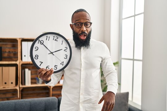 African american man holding big clock at the office scared and amazed with open mouth for surprise, disbelief face