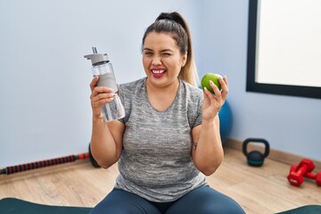 Young hispanic woman holding green apple and bottle of water winking looking at the camera with...