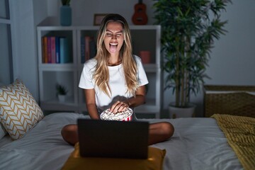 Young blonde woman sitting on the bed at home watching a movie from laptop sticking tongue out...