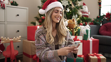 Obraz na płótnie Canvas Young blonde woman using smartphone sitting by christmas tree at home