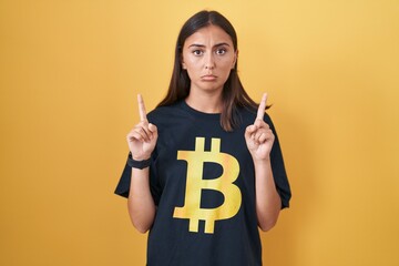 Young hispanic woman wearing bitcoin t shirt pointing up looking sad and upset, indicating...