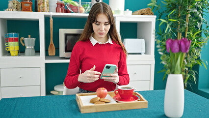 Young hispanic woman having breakfast using smartphone at dinning room