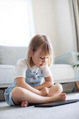 A cute girl looks at the tablet screen while sitting on the floor in the room. Children's educational apps for tablet. 