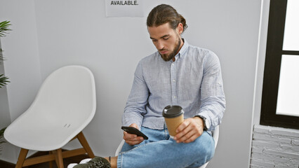 Young hispanic man using smartphone drinking coffee at waiting room