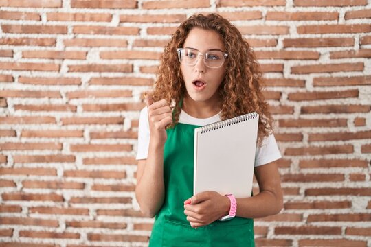 Young caucasian woman holding art notebook surprised pointing with hand finger to the side, open mouth amazed expression.