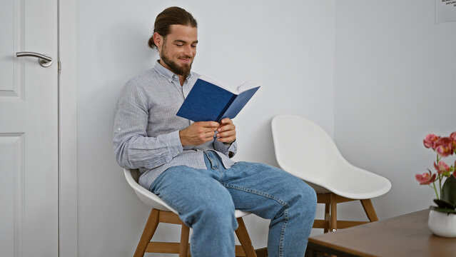 Young hispanic man reading book sitting on chair smiling at waiting room