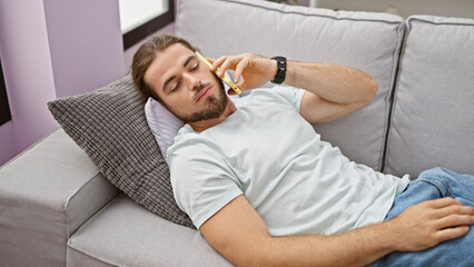 Young hispanic man talking on smartphone lying on sofa at home