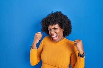 Fototapeta na wymiar Black woman with curly hair standing over blue background very happy and excited doing winner gesture with arms raised, smiling and screaming for success. celebration concept.