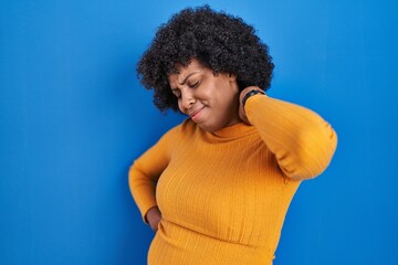 Black woman with curly hair standing over blue background suffering of neck ache injury, touching neck with hand, muscular pain