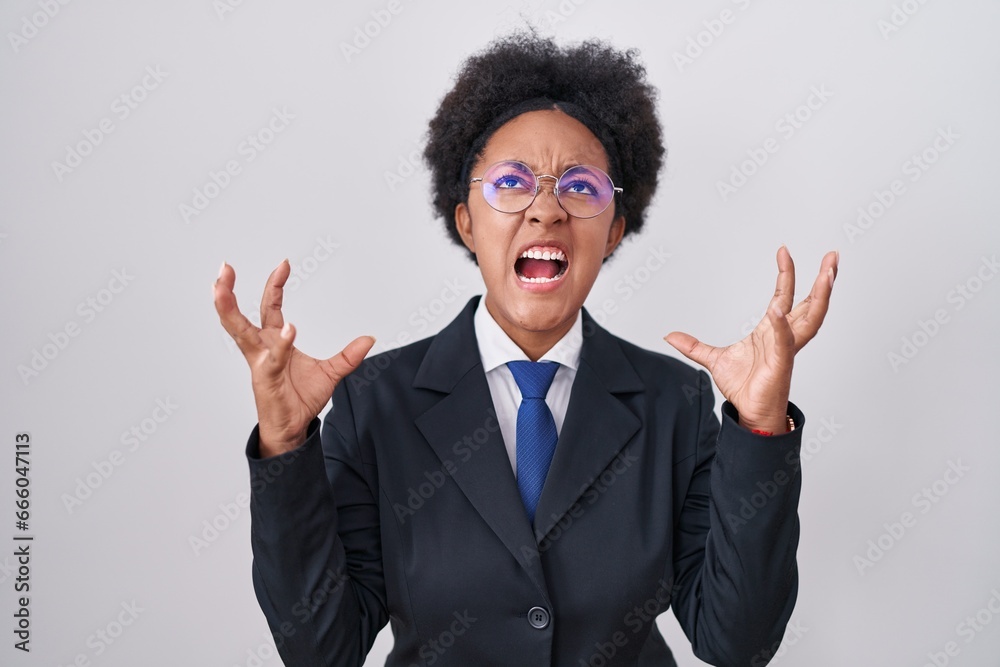 Sticker Beautiful african woman with curly hair wearing business jacket and glasses crazy and mad shouting and yelling with aggressive expression and arms raised. frustration concept.