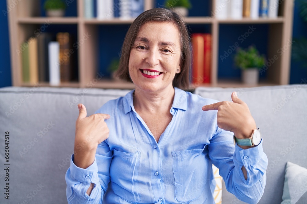 Wall mural Middle age hispanic woman sitting on the sofa at home looking confident with smile on face, pointing oneself with fingers proud and happy.