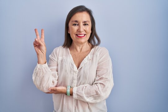 Middle age hispanic woman standing over blue background smiling with happy face winking at the camera doing victory sign with fingers. number two.